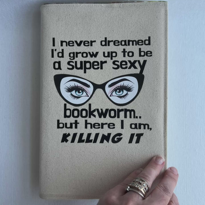 a short video  showing  the inside of a book covered in a reusable linen-color canvas book cover, with the words, 'I never dreamed I'd grow up to be a super sexy bookworm, but here I am killing it.' A pair of eyes with long lashes behind cat eye glasses completes the design.