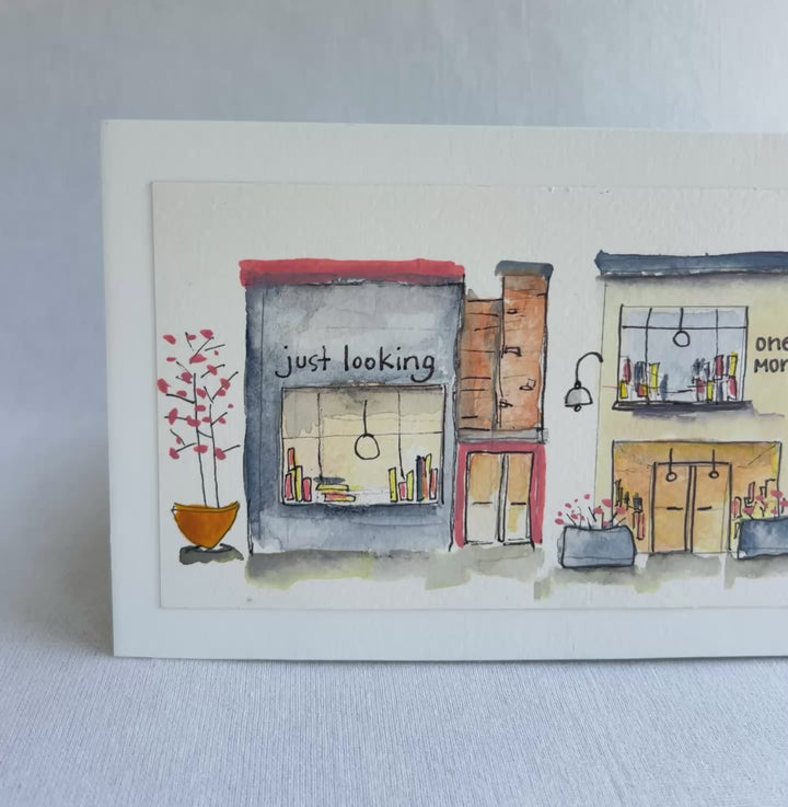 A watercolor painting of four colorful brick storefront bookstores, with the names, "Just Looking"  "One More" and "Last Chapter."  Displayed on top of a flower-painted book.
