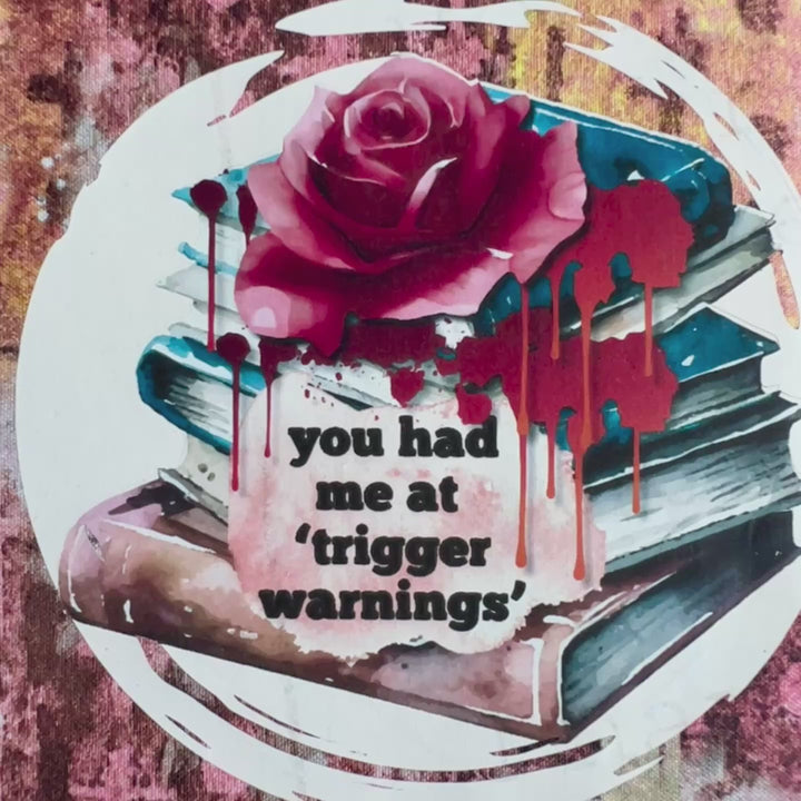 a short video showing a book covered in a colorful fabric reusable book cover in colors of pinks and orange. A white circle on the front contains an original design with a stack of books, a rose, blood drips and the words, 'you had me at trigger warnings' finishes the look.  Meant for readers of dark romance.