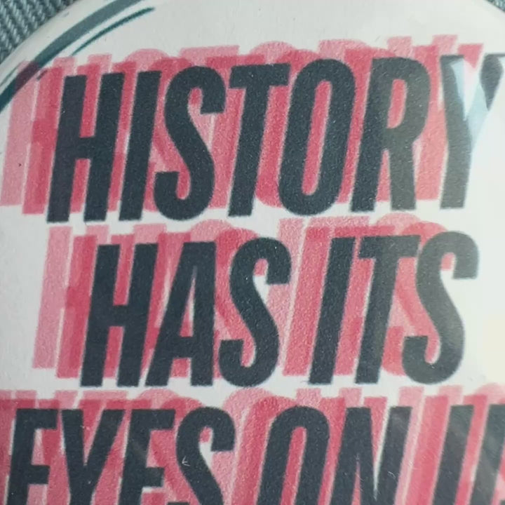 a short video of a round pinback button with a black background, yellow and red flames surrounding the words, "tenth circle added to rapidly growing hell." A second button with a yellow background and a cartoon image of an angry book has the words, 'ban fascists not books.' A third button has a white background and the words, 'history has its eyes on us' in blue and red.  All pinned to a jean jacket for display.