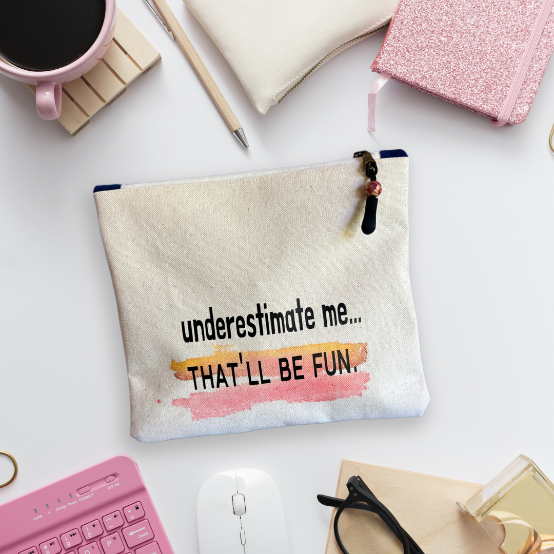 a canvas zip bag with the words, "underestimate me, that'll be fun" in black lettering. the bag has watercolor painting on it, and is surrounded by office tools