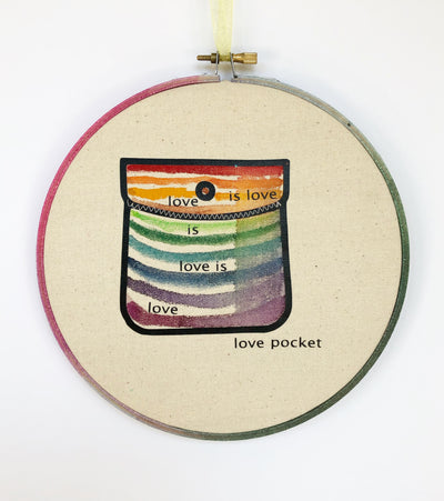 round wooden hoop with canvas and a multicolored striped painted pocket with the words, "love is love is love...love pocket."