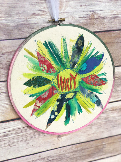 round dyed wooden hoop art, with painted and stitched fabric leaves in colors of yellow and orange and red, with the word 'happy' painted in the center