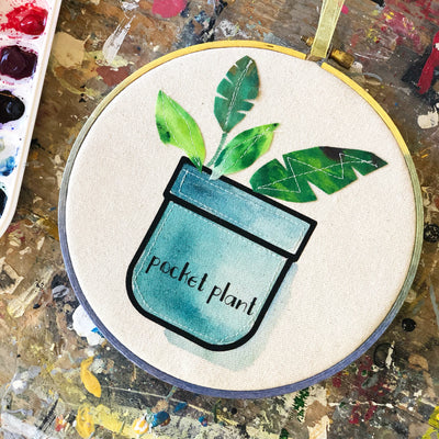 round wooden painted hoop with canvas, a painted blue pocket with plant leaves, and the words, 'pocket plant.'