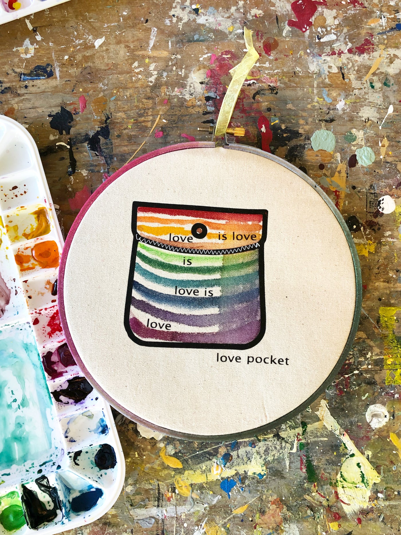 round wooden hoop with canvas and a multicolored striped painted pocket with the words, "love is love is love...love pocket."