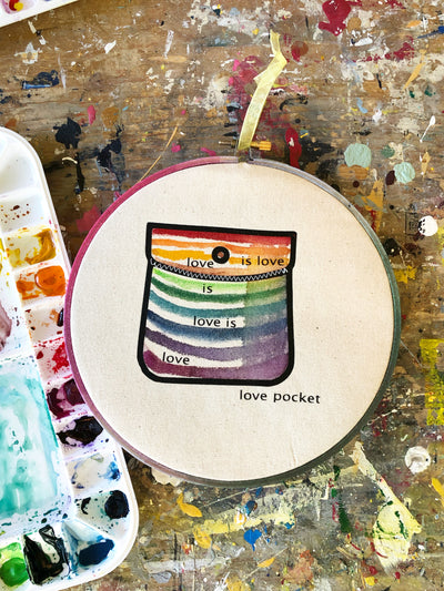 round wooden hoop with canvas and a multicolored striped painted pocket with the words, "love is love is love...love pocket."