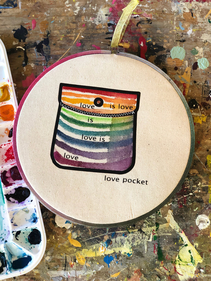 round wooden hoop with canvas and a multicolored striped painted pocket with the words, "love is love is love...love pocket."