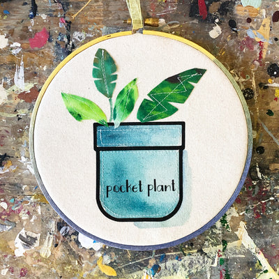 round wooden painted hoop with canvas, a painted blue pocket with plant leaves, and the words, 'pocket plant.'