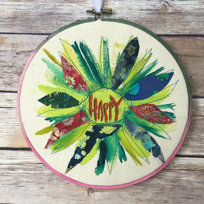 round dyed wooden hoop art, with painted and stitched fabric leaves in colors of yellow and orange and red, with the word 'happy' painted in the center