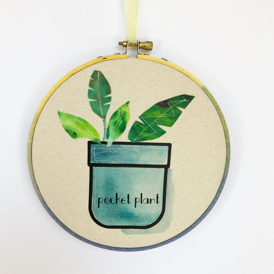 round wooden painted hoop with canvas, a painted blue pocket with plant leaves, and the words, 'pocket plant.'