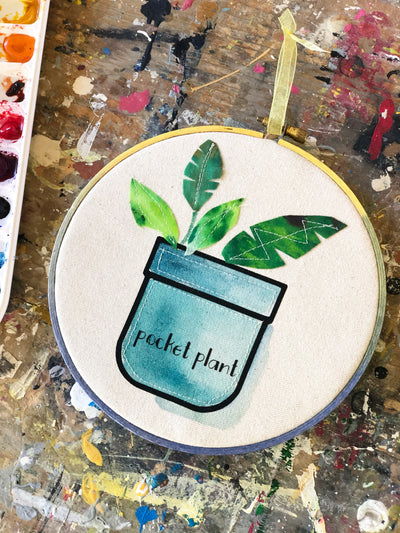 round wooden painted hoop with canvas, a painted blue pocket with plant leaves, and the words, 'pocket plant.'