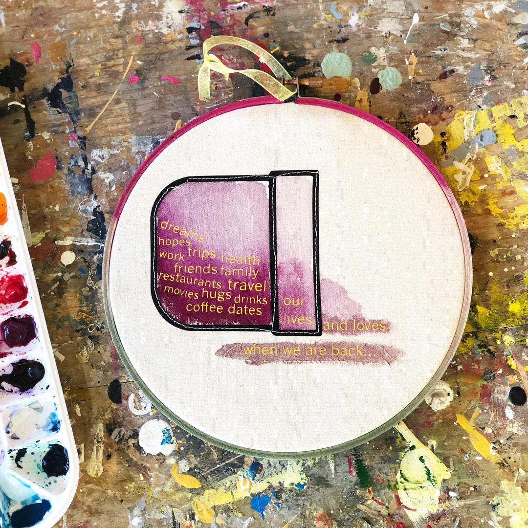round wooden painted hoop with canvas and a painted pocket on its side, with the words "dreams, hopes, trips, health, friends, family, travel, hugs, drinks, coffee dates, our lives and loves" spilling out.