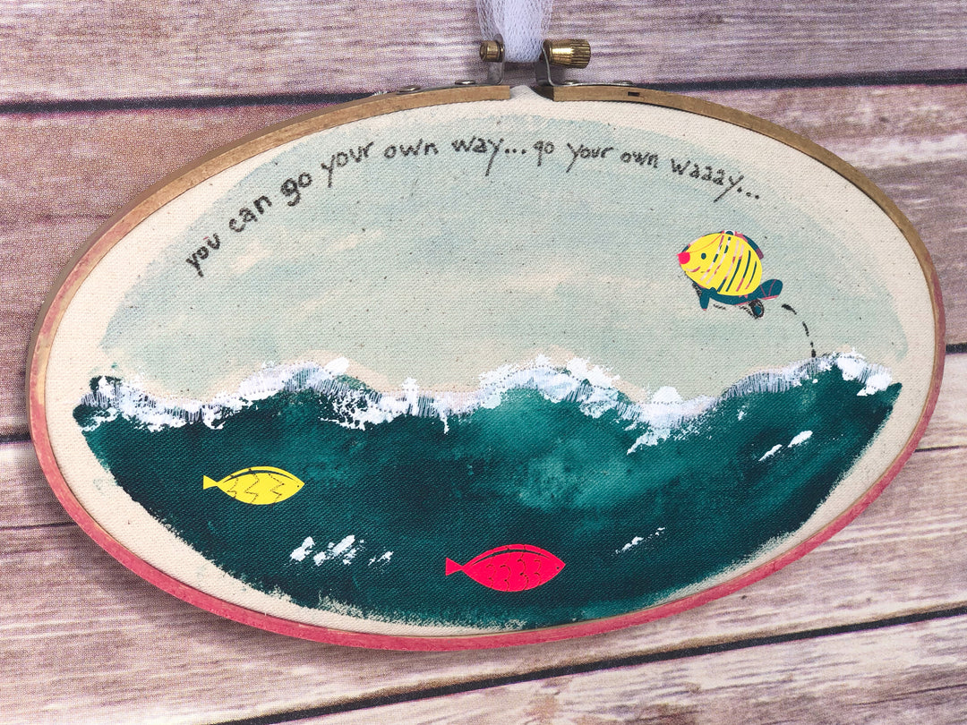 oval wooden dyed hoop art with fabric painted to look like the seashore, with jumping fish and the words, "you can go your own way.."