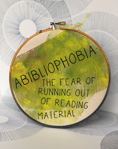 round wooden hoop with yellow-green paint and the words, "Abibliophobia - the fear of running out of reading material."