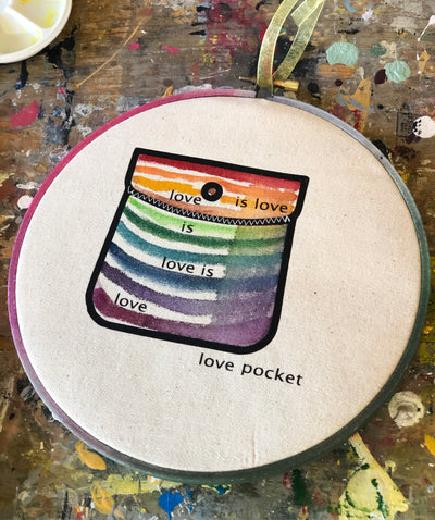 round wooden hoop with canvas and a multicolored striped painted pocket with the words, "love is love is love...love pocket."