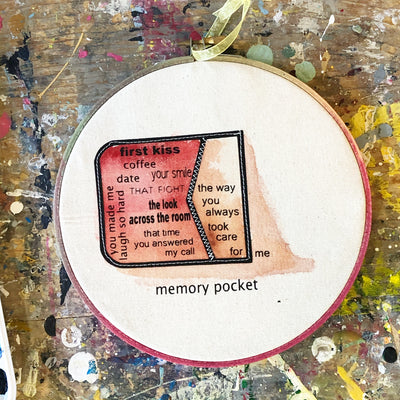 a round wooden painted hoop with canvas, a painted red pocket on its side with the words spilling out...memory pocket, first kiss, coffee date, your smile, the look across the room, that time you answered my call, you always made me laugh so har, the way you always took care for me.