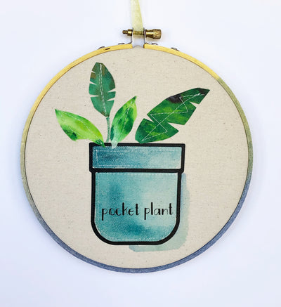 round wooden painted hoop with canvas, a painted blue pocket with plant leaves, and the words, 'pocket plant.'