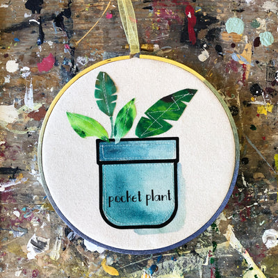 round wooden painted hoop with canvas, a painted blue pocket with plant leaves, and the words, 'pocket plant.'