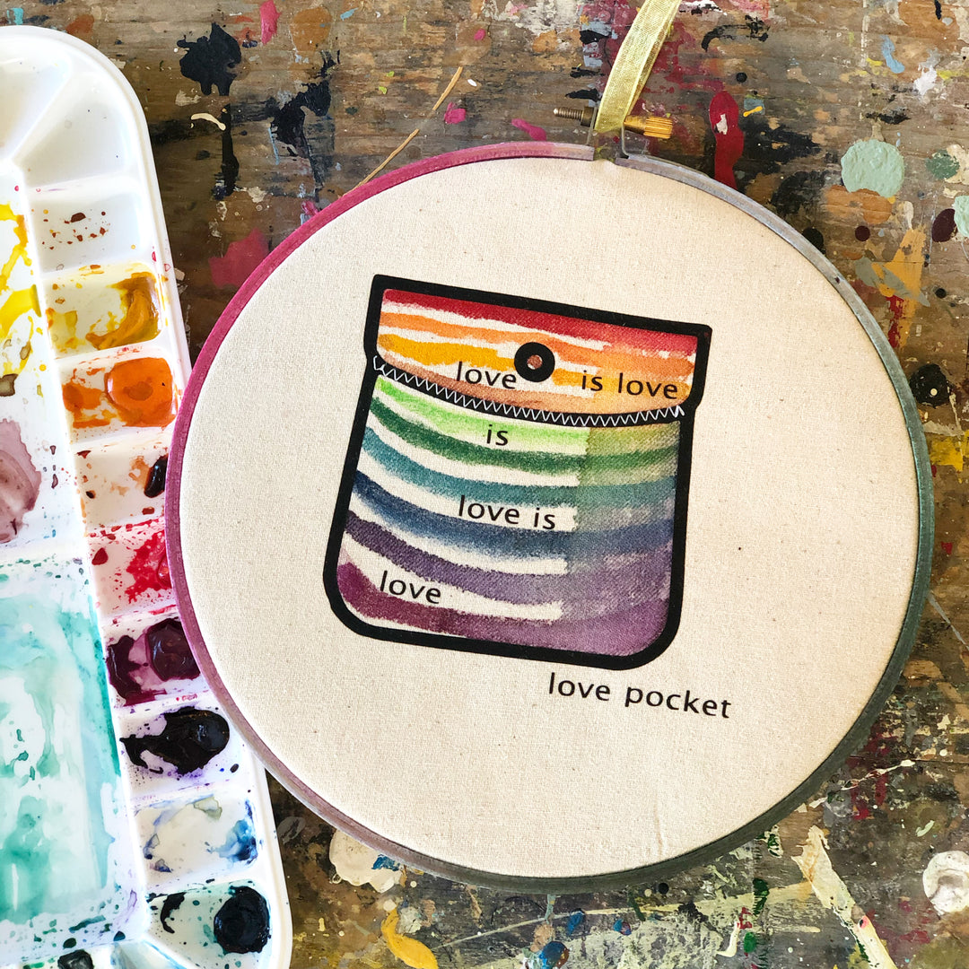 round wooden hoop with canvas and a multicolored striped painted pocket with the  words, "love is love is love...love pocket."