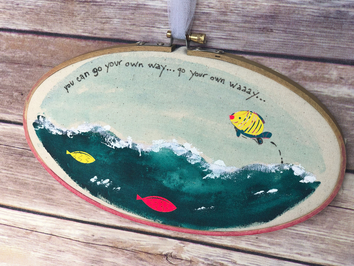 oval wooden dyed hoop art with fabric painted to look like the seashore, with jumping fish and the words, "you can go your own way.."