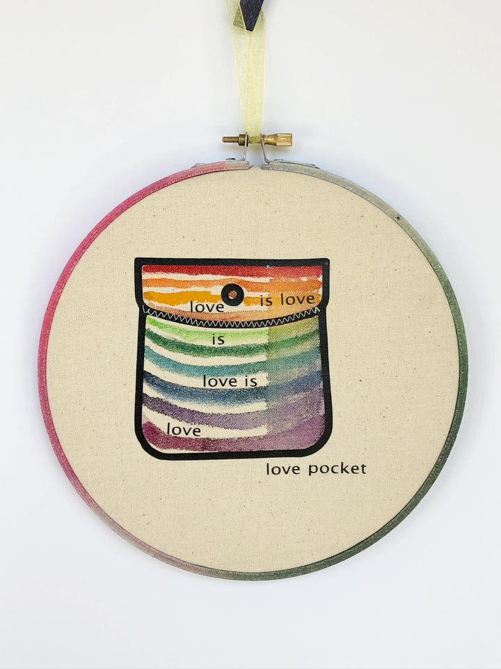 round wooden hoop with canvas and a multicolored striped painted pocket with the words, "love is love is love...love pocket."