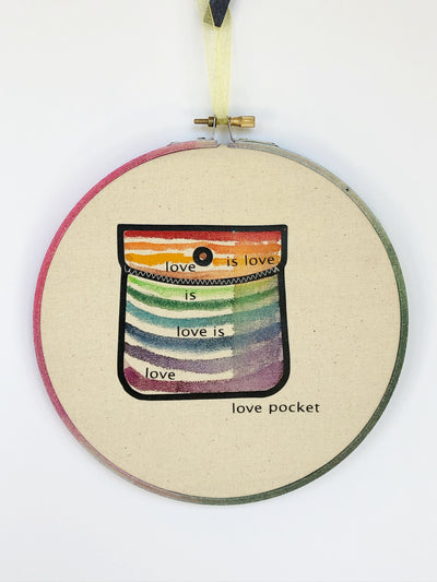 round wooden hoop with canvas and a multicolored striped painted pocket with the words, "love is love is love...love pocket."