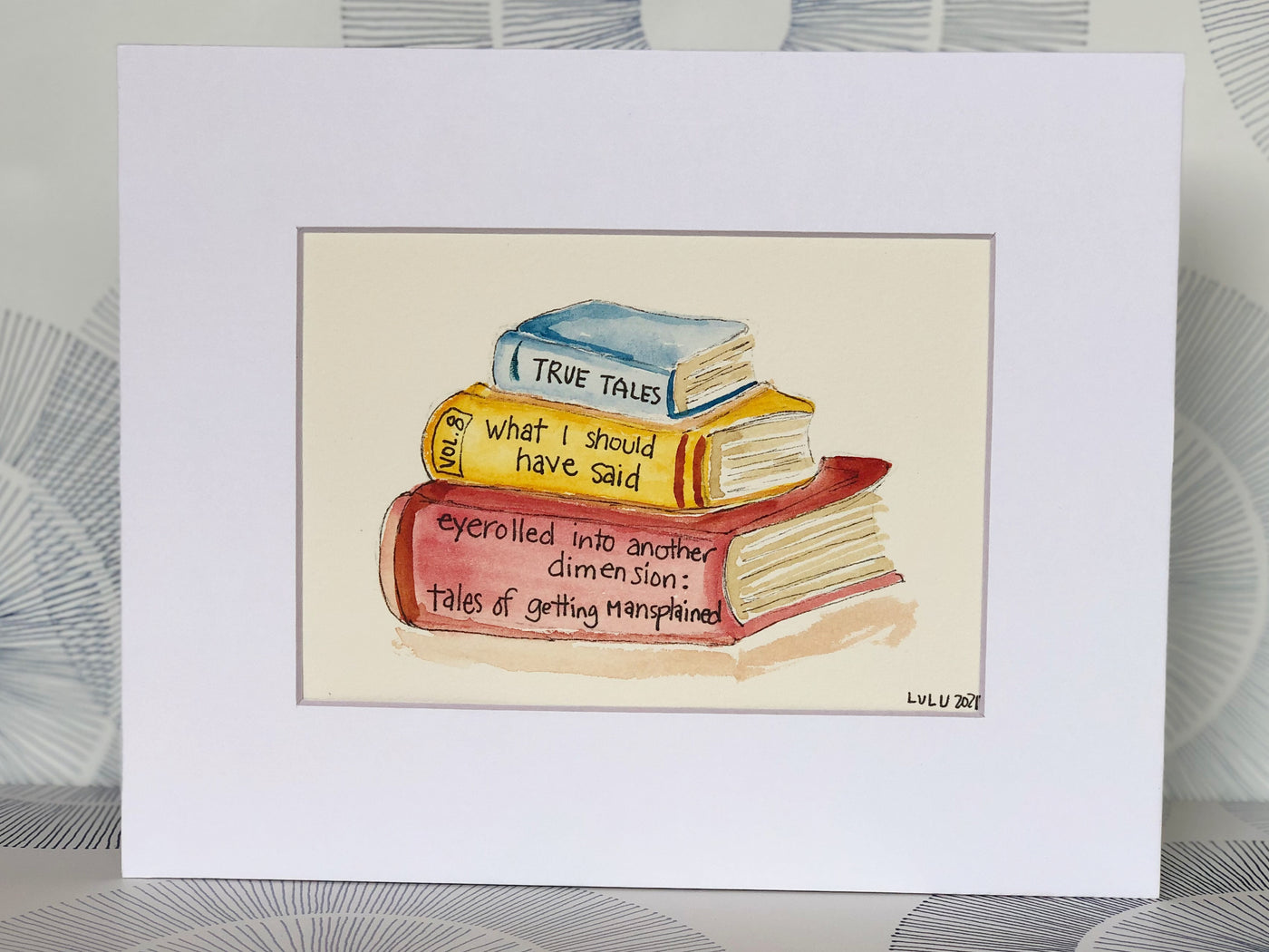 an original painted watercolor piece with three books in colors of burnt yellow, brick red and faded cerulean blue