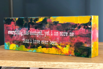 a painted wooden panel, with paint colors of teal, burnt yellow, red, coral and black, with the words, "everything has changed...yet I am more me than i have ever been."