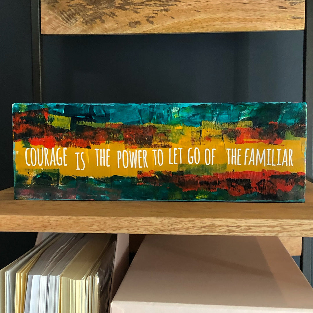 a painted wooden panel, with paint colors of teal, burnt yellow, red, coral and black, with the words, "courage is the power to let go of the familiar."
