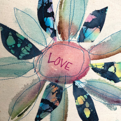round dyed wooden hoop art, with painted and stitched fabric leaves in colors of blue and pink and green, with the word 'love' painted in the center