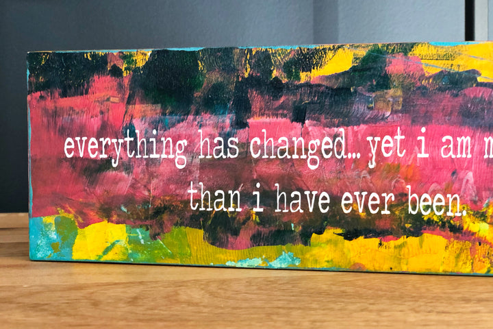 a painted wooden panel, with paint colors of teal, burnt yellow, red, coral and black, with the words, "everything has changed...yet I am more me than i have ever been."