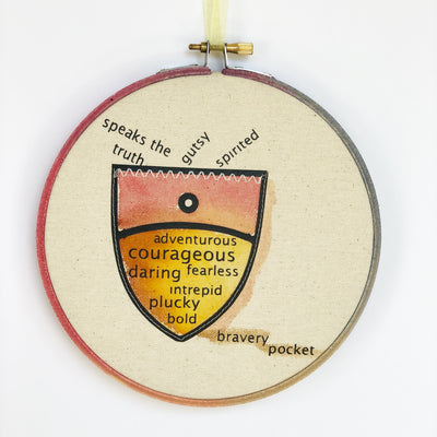 round painted wooden hoop with canvas and a painted pocket shape, filled with words about bravery, such as speaks the truth, gutsy, spirited, adventurous, courageous, fearless, daring, intrepid, plucky and bold