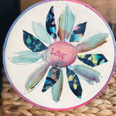 round dyed wooden hoop art, with painted and stitched fabric leaves in colors of blue and pink and green, with the word 'love' painted in the center