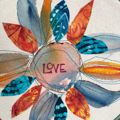 round dyed wooden hoop art, with painted and stitched fabric leaves in colors of blue and pink and green, with the word 'love' painted in the center