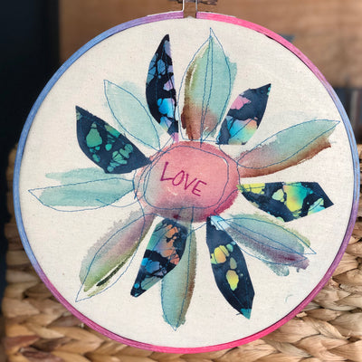 round dyed wooden hoop art, with painted and stitched fabric leaves in colors of blue and pink and green, with the word 'love' painted in the center
