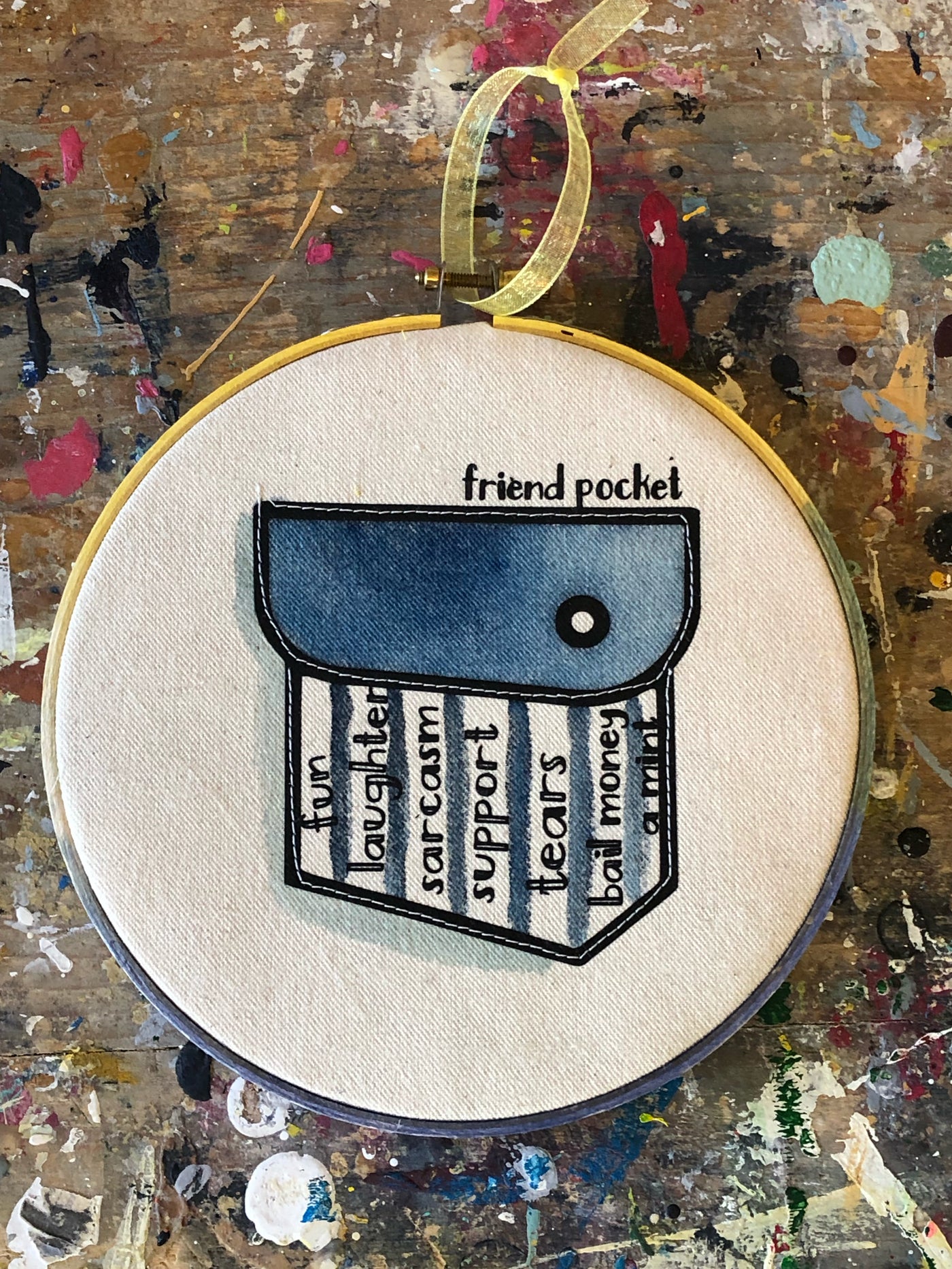 round painted wooden hoop with canvas and a painted blue pocket, containing the words 'friend pocket' and the words, fun, laughter, sarcasm, support, tears, bail money, a mint.'