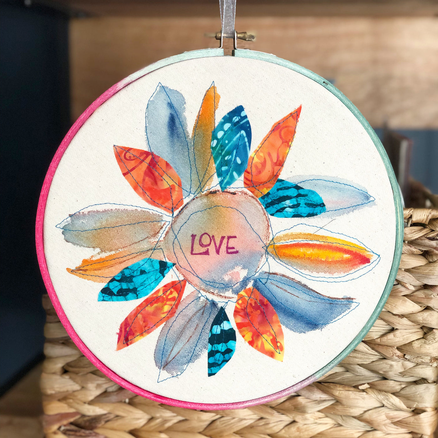 round dyed wooden hoop art, with painted and stitched fabric leaves in colors of blue and pink and green, with the word 'love' painted in the center