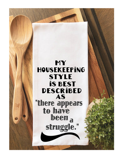 a white cotton kitchen towel with the words, "my housekeeping style is best described as "there appears to have been a struggle."